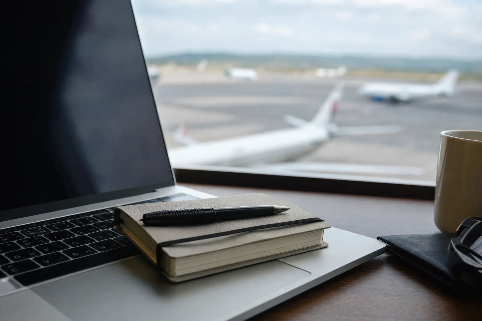 A small notebook and pen resting on a laptop keyboard in an airport lounge, with a blurred runway and parked aircraft visible through the window behind